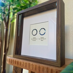 May include: A framed print displaying two watch bezel designs, one blue and one black, with the text "Rolex GMT-Master II" and model names. The dark wood frame surrounds a white mount, creating a striking contrast.