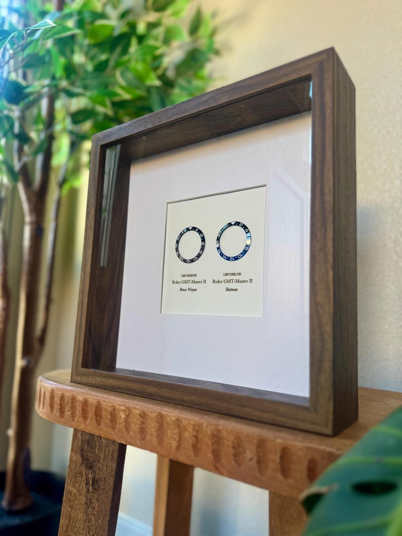 May include: A framed print displaying two watch bezel designs, one blue and one black, with the text "Rolex GMT-Master II" and model names. The dark wood frame surrounds a white mount, creating a striking contrast.