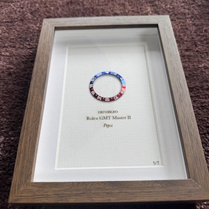 May include: Framed artwork featuring a watch bezel with red and blue coloring, set against a white background. The frame is a dark brown wood. Text on the artwork reads "126710BLRO Rolex GMT Master II Pepsi" and "D.T."