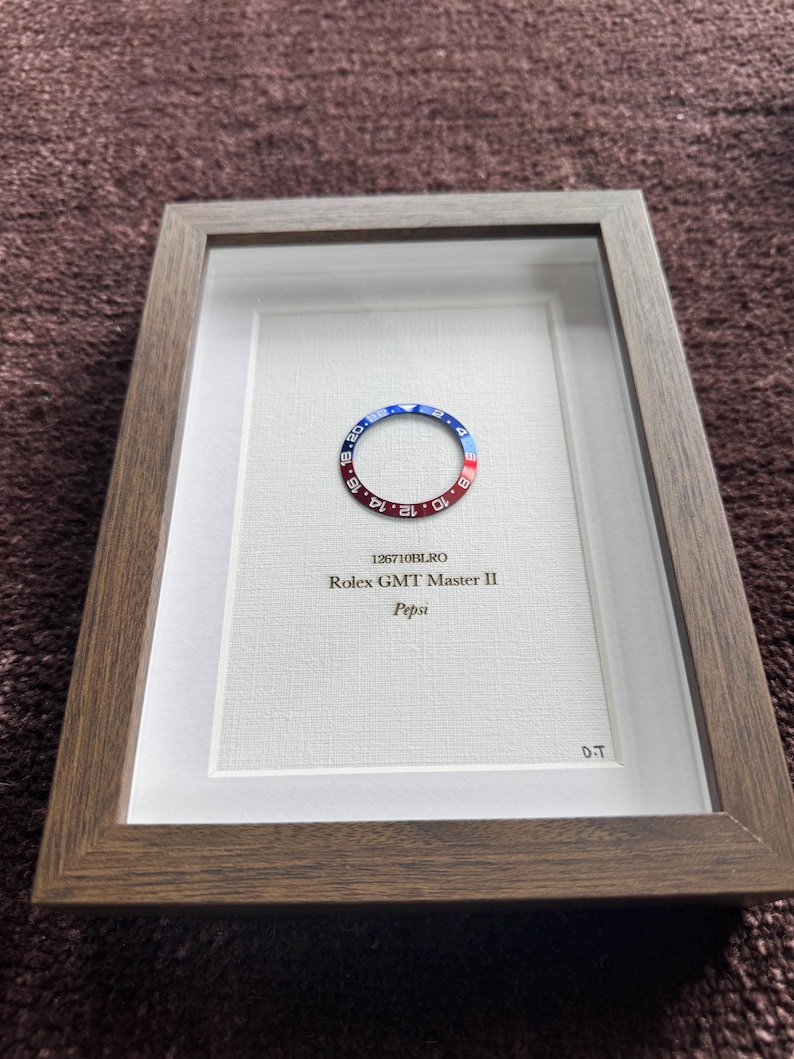 May include: Framed artwork featuring a watch bezel with red and blue coloring, set against a white background. The frame is a dark brown wood. Text on the artwork reads "126710BLRO Rolex GMT Master II Pepsi" and "D.T."
