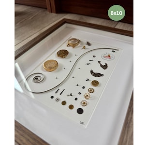 Puede incluir: Una obra de arte enmarcada que presenta componentes de reloj sobre un fondo blanco. La disposici&oacute;n incluye engranajes, resortes y carcasas de reloj en tonos dorados y plateados. El marco es de madera marr&oacute;n claro y la obra est&aacute; etiquetada como "8x10".