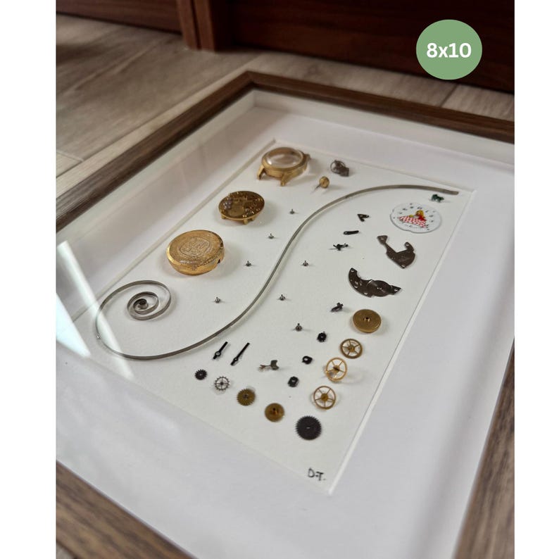 Puede incluir: Una obra de arte enmarcada que presenta componentes de reloj sobre un fondo blanco. La disposici&oacute;n incluye engranajes, resortes y carcasas de reloj en tonos dorados y plateados. El marco es de madera marr&oacute;n claro y la obra est&aacute; etiquetada como "8x10".