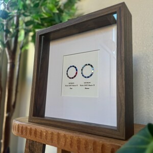 May include: A dark brown framed print displaying two watch bezels with red, blue, and black designs. The frame is made of wood and is placed on a wooden stool. The print includes the text "Rolex GMT-Master II" and bezel style names.