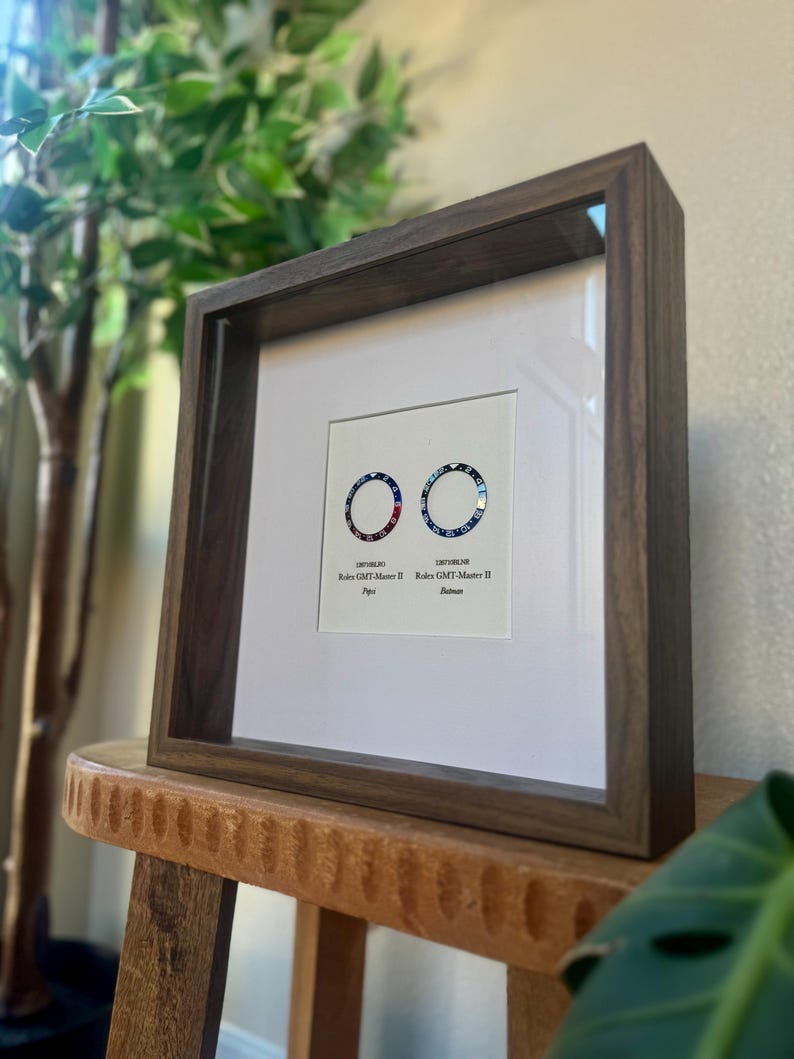 May include: A dark brown framed print displaying two watch bezels with red, blue, and black designs. The frame is made of wood and is placed on a wooden stool. The print includes the text "Rolex GMT-Master II" and bezel style names.