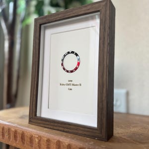 May include: A framed print featuring a watch bezel design with red and black segments. The print includes the text "16760 Rolex GMT-Master II Coke" in a minimalist style. The frame is a dark brown color.