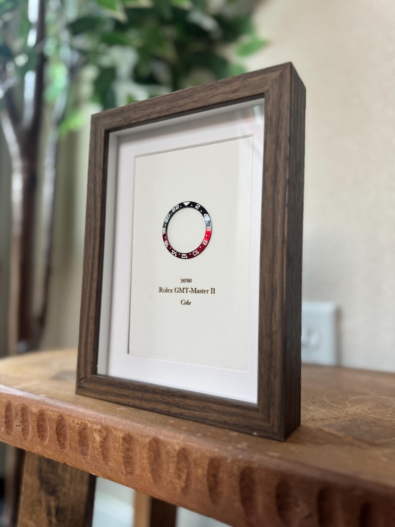May include: A framed print featuring a watch bezel design with red and black segments. The print includes the text "16760 Rolex GMT-Master II Coke" in a minimalist style. The frame is a dark brown color.
