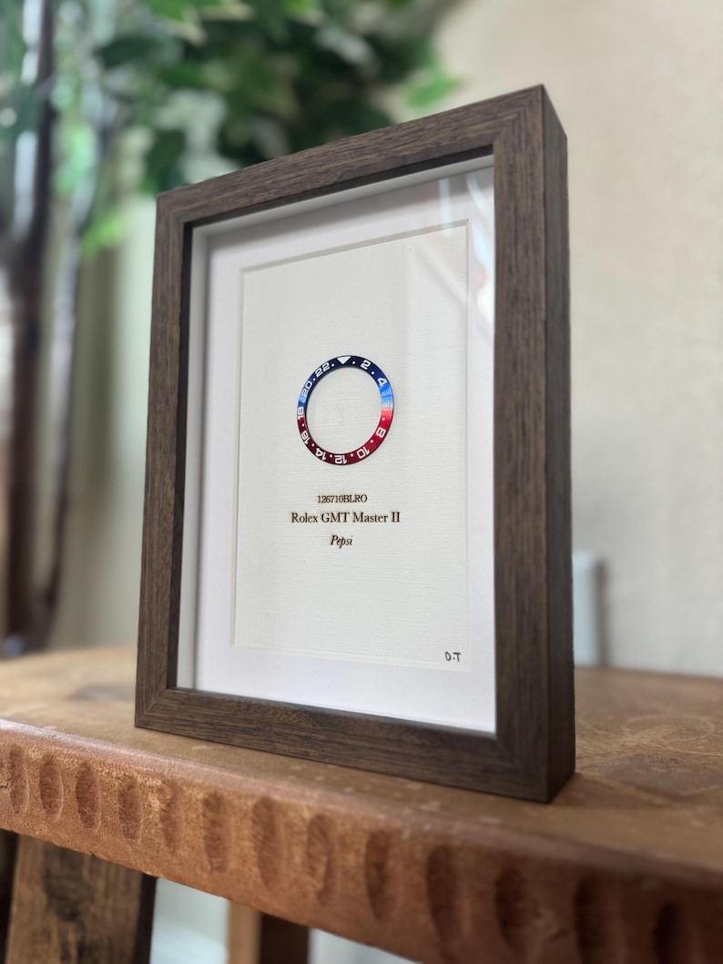 May include: Framed print featuring a Rolex GMT Master II "Pepsi" bezel design. The bezel is red and blue, with white text and numbers. The frame is dark brown, and the print includes the model number and "Pepsi" text.