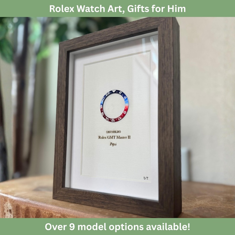 May include: Framed artwork featuring a Rolex GMT Master II "Pepsi" watch bezel. The bezel is red and blue, with white numerals. The artwork is in a dark brown wooden frame, with the text "126710BLRO" and "Pepsi" visible.