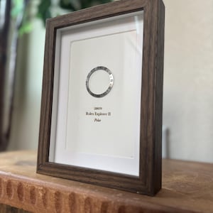 May include: A framed print featuring a watch bezel design with the text "Rolex Explorer II Polar." The frame is a dark brown color, and the print is set against a white background. The print is displayed on a wooden surface.