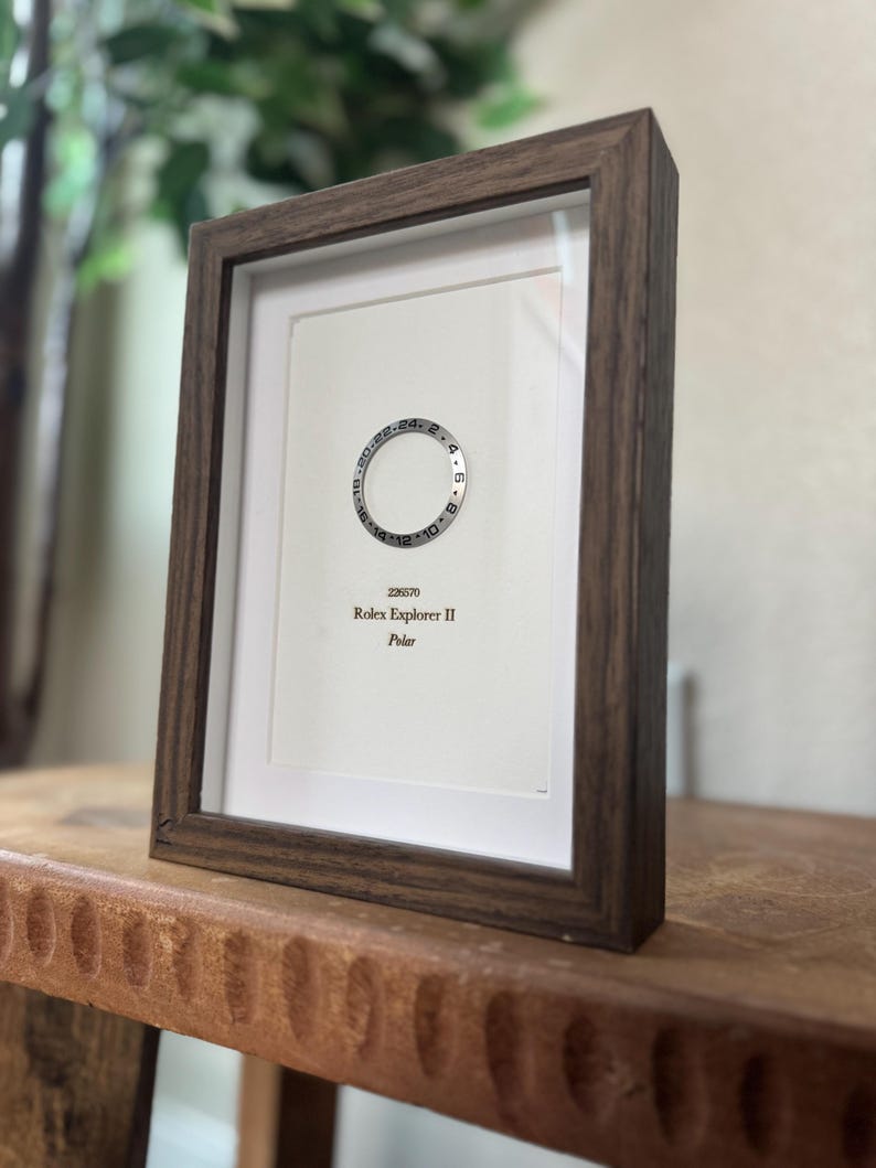 May include: A framed print featuring a watch bezel design with the text "Rolex Explorer II Polar." The frame is a dark brown color, and the print is set against a white background. The print is displayed on a wooden surface.