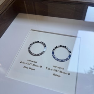 May include: A framed display showcasing two Rolex GMT-Master II watch bezels. One is black and grey, the other blue and black. Text below reads "126710GRNR Rolex GMT-Master II Bruce Wayne" and "126710BLNR Rolex GMT-Master II Batman."
