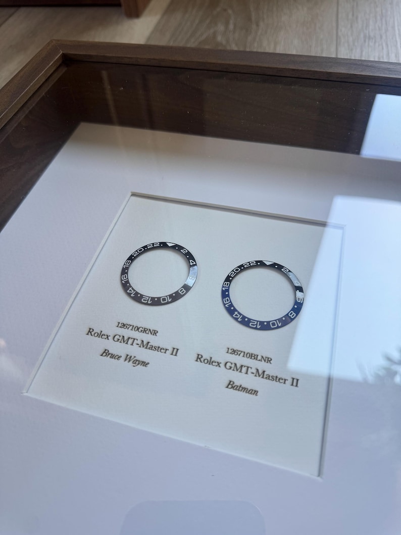 May include: A framed display showcasing two Rolex GMT-Master II watch bezels. One is black and grey, the other blue and black. Text below reads "126710GRNR Rolex GMT-Master II Bruce Wayne" and "126710BLNR Rolex GMT-Master II Batman."