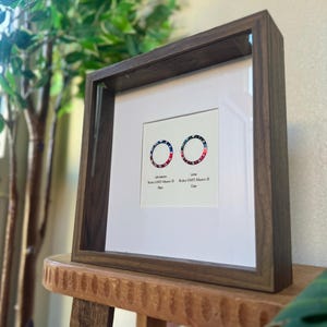May include: A framed print with two circular designs in red, white, and blue, reminiscent of watch bezels. The dark brown wooden frame sits on a wooden stool. The text "Rolex GMT-Master II" is displayed below the designs.