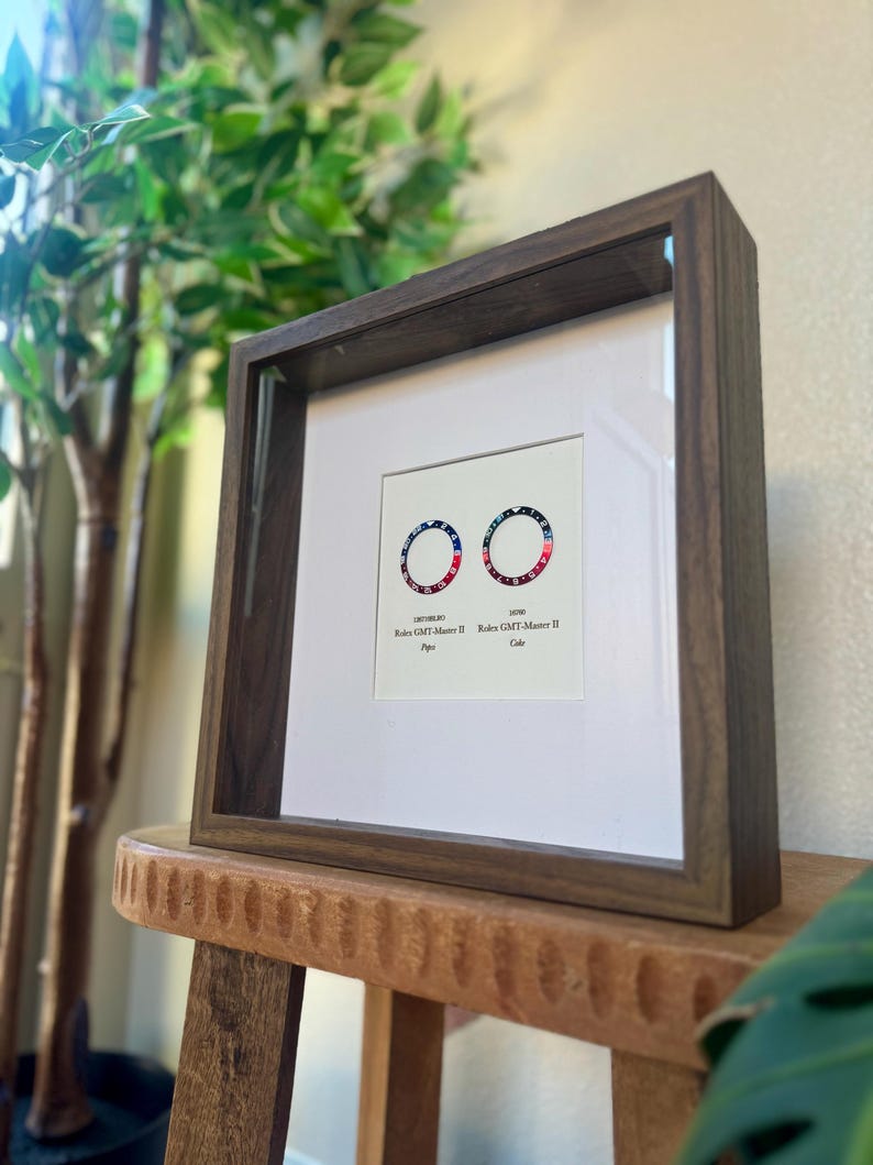 May include: A framed print with two circular designs in red, white, and blue, reminiscent of watch bezels. The dark brown wooden frame sits on a wooden stool. The text "Rolex GMT-Master II" is displayed below the designs.