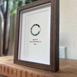 May include: A framed artwork featuring a Rolex GMT Master II Sprite bezel. The black and silver bezel is set against a white background, encased in a dark brown wooden frame. The text "126720VTNR Rolex GMT Master II Sprite" is printed below the bezel.
