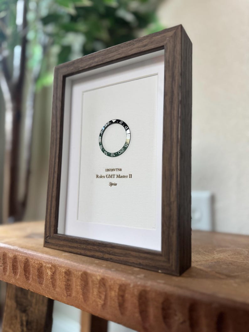 May include: A framed artwork featuring a Rolex GMT Master II Sprite bezel. The black and silver bezel is set against a white background, encased in a dark brown wooden frame. The text "126720VTNR Rolex GMT Master II Sprite" is printed below the bezel.