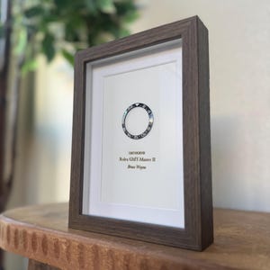 May include: A framed print featuring a watch bezel design. The print includes the text "126710GRNR Rolex GMT-Master II Bruce Wayne" in a dark brown wooden frame. The background is white.