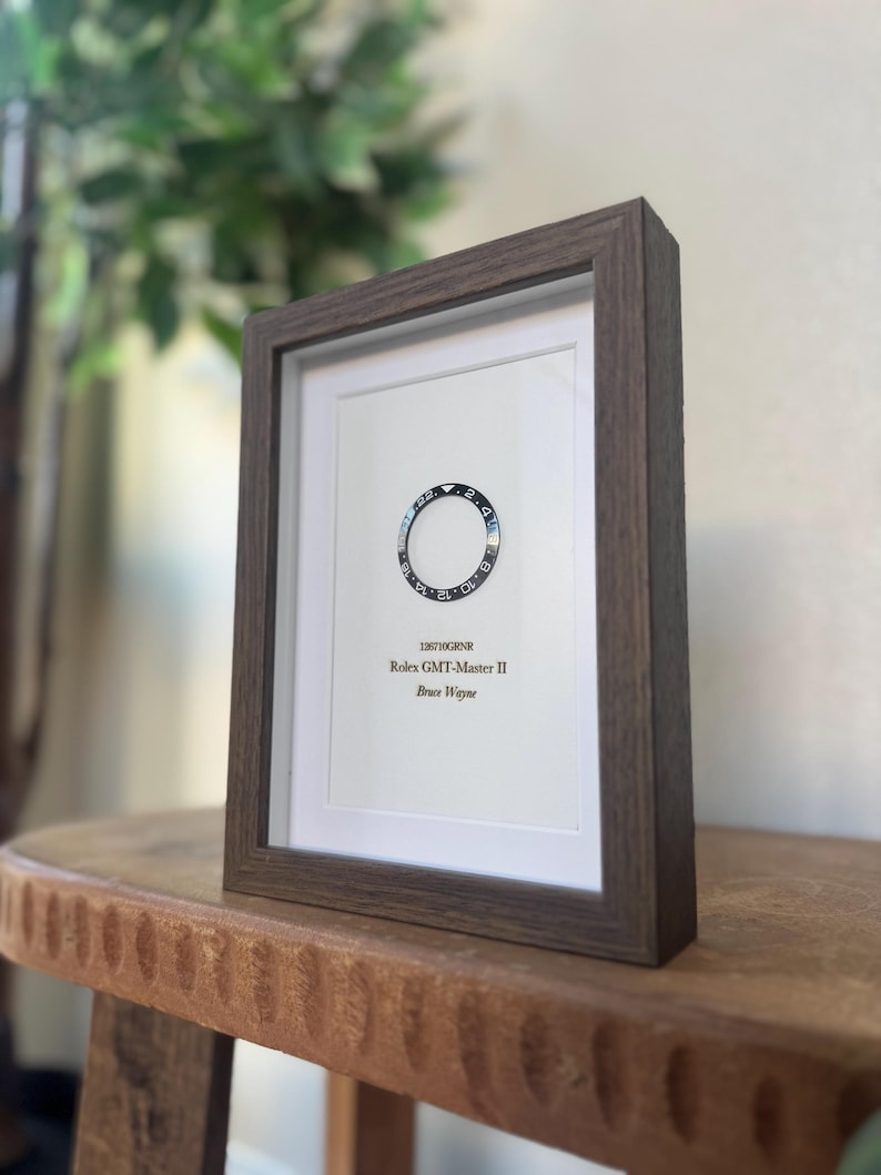 May include: A framed print featuring a watch bezel design. The print includes the text "126710GRNR Rolex GMT-Master II Bruce Wayne" in a dark brown wooden frame. The background is white.