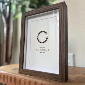 May include: A framed print featuring a Rolex GMT-Master II Root Beer bezel. The bezel is black and gold, set against a white background. The frame is a dark brown wood, and the print includes the text "126711CHNR".