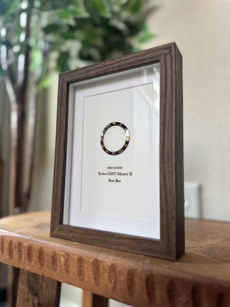 May include: A framed print featuring a Rolex GMT-Master II Root Beer bezel. The bezel is black and gold, set against a white background. The frame is a dark brown wood, and the print includes the text "126711CHNR".