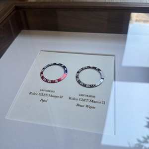 May include: A framed display featuring two Rolex GMT-Master II watch bezels. One is red and blue, the "Pepsi" model, and the other is black and grey, the "Bruce Wayne" model. Both bezels are circular with numerical markings.