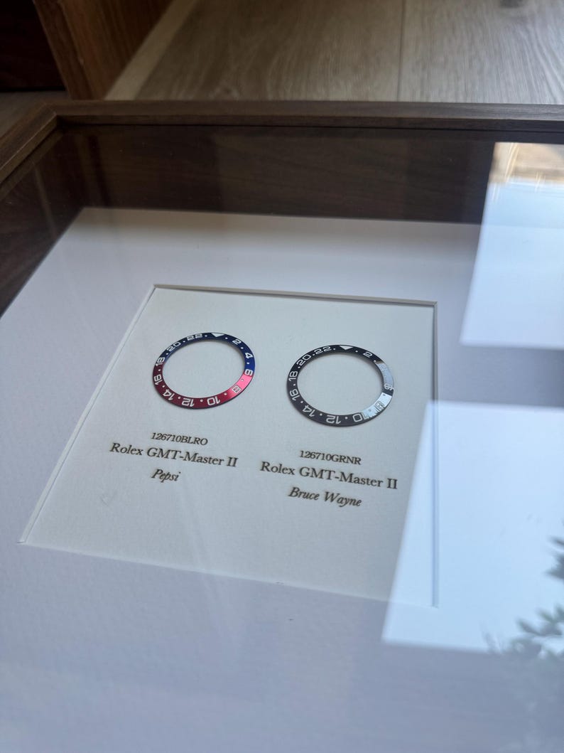May include: A framed display featuring two Rolex GMT-Master II watch bezels. One is red and blue, the "Pepsi" model, and the other is black and grey, the "Bruce Wayne" model. Both bezels are circular with numerical markings.
