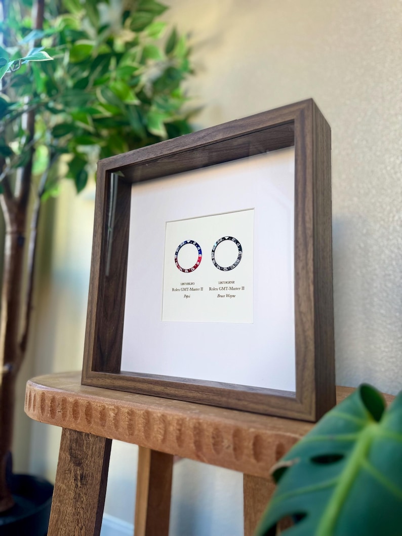 May include: A framed print with two circular designs in red, white, and blue, resembling watch faces. The dark wood frame surrounds a white mount. Text is visible on the print. The artwork is displayed on a wooden stool.