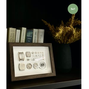 Puede incluir: Una obra de arte enmarcada de 5x7 pulgadas muestra componentes de reloj, incluyendo esferas y engranajes, sobre un fondo blanco. El marco marr&oacute;n oscuro est&aacute; sobre una estanter&iacute;a con libros y un jarr&oacute;n negro con flores secas.