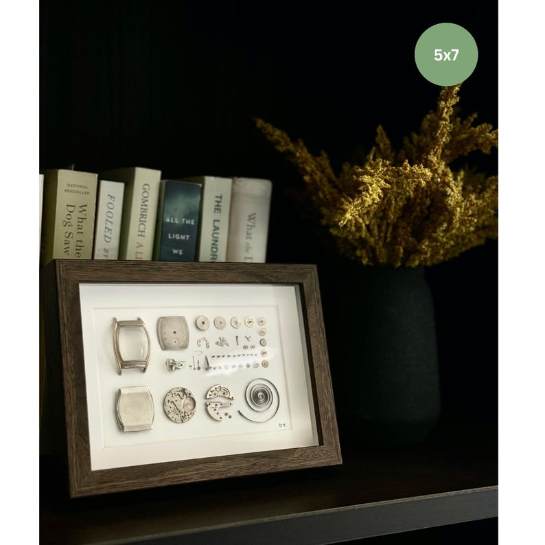 Puede incluir: Una obra de arte enmarcada de 5x7 pulgadas muestra componentes de reloj, incluyendo esferas y engranajes, sobre un fondo blanco. El marco marr&oacute;n oscuro est&aacute; sobre una estanter&iacute;a con libros y un jarr&oacute;n negro con flores secas.