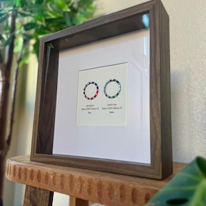 May include: A framed print showing two watch bezel designs, one with red, white, and blue accents, the other with blue and green. The dark wood frame surrounds a white mount, displaying the watch designs.