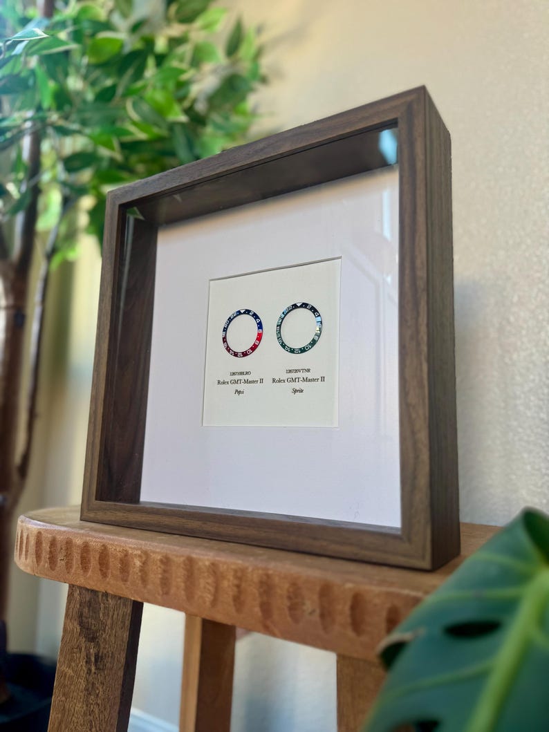 May include: A framed print showing two watch bezel designs, one with red, white, and blue accents, the other with blue and green. The dark wood frame surrounds a white mount, displaying the watch designs.