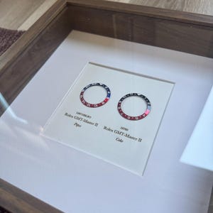 May include: Two Rolex GMT-Master II bezels, one with a red and blue Pepsi design, the other with a black and red Coke design, presented in a framed display. The text "Pepsi" and "Coke" are visible.