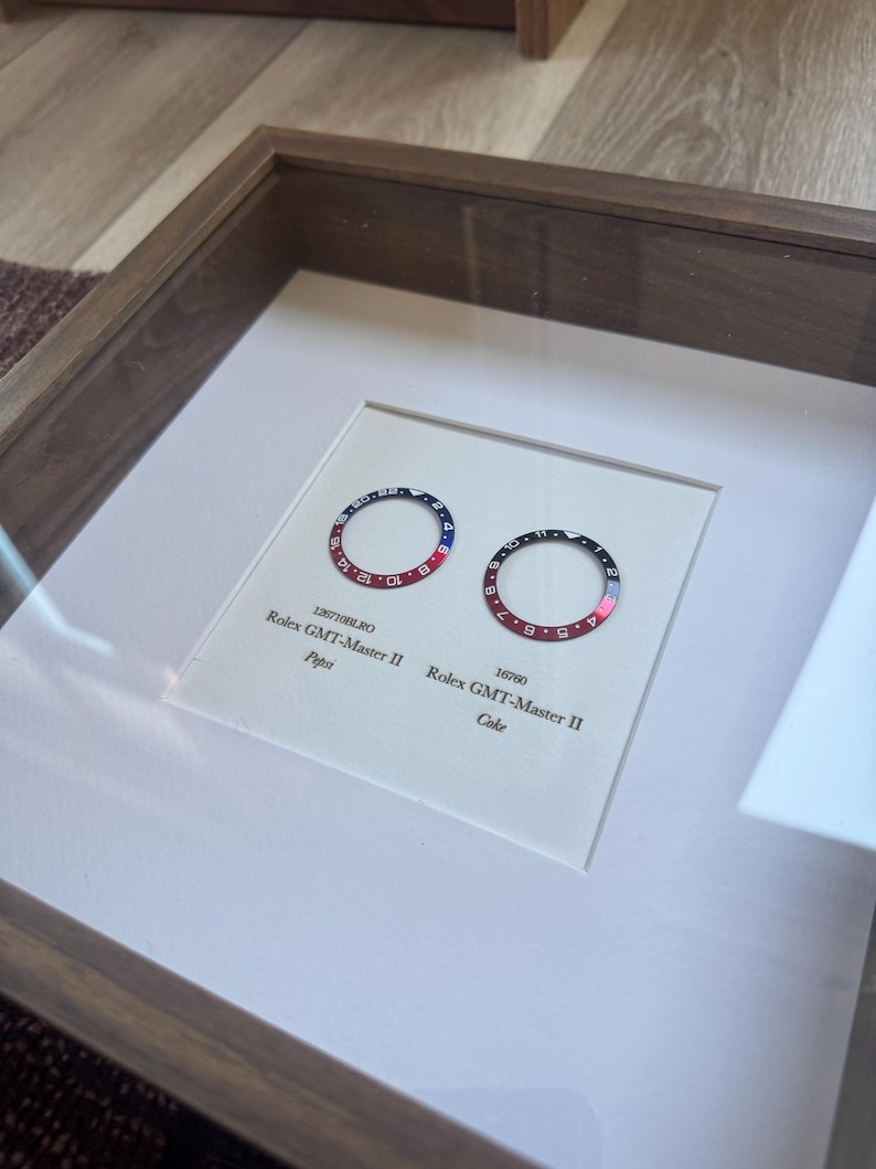 May include: Two Rolex GMT-Master II bezels, one with a red and blue Pepsi design, the other with a black and red Coke design, presented in a framed display. The text "Pepsi" and "Coke" are visible.