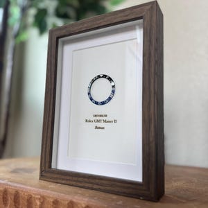 May include: Framed artwork featuring a Rolex GMT Master II Batman watch bezel. The bezel is black and blue, set against a white background. The frame is made of dark brown wood. Text on the artwork reads "126710BLNR Rolex GMT Master II Batman."
