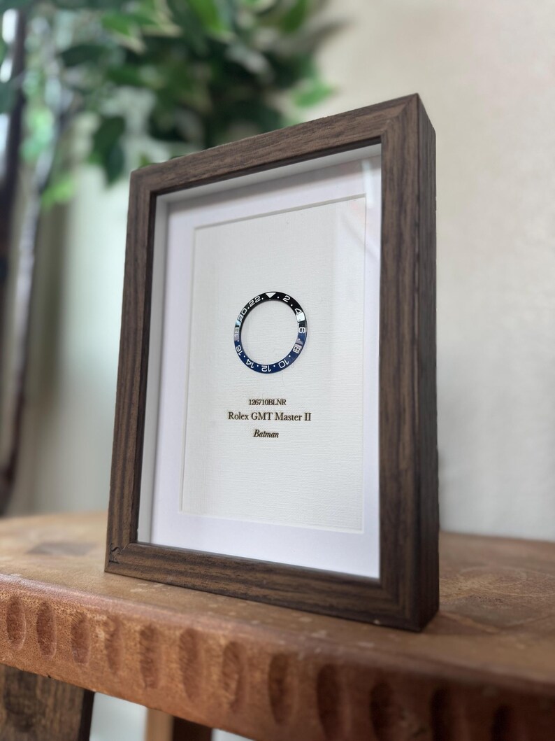 May include: Framed artwork featuring a Rolex GMT Master II Batman watch bezel. The bezel is black and blue, set against a white background. The frame is made of dark brown wood. Text on the artwork reads "126710BLNR Rolex GMT Master II Batman."