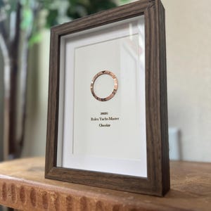 May include: Framed print featuring a rose gold watch bezel with the text "Rolex Yacht-Master Chocolate." The print is set in a dark brown wooden frame, displayed on a wooden surface.
