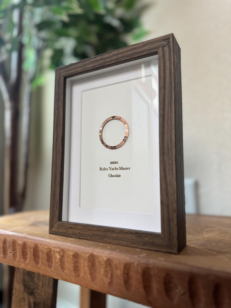 May include: Framed print featuring a rose gold watch bezel with the text "Rolex Yacht-Master Chocolate." The print is set in a dark brown wooden frame, displayed on a wooden surface.