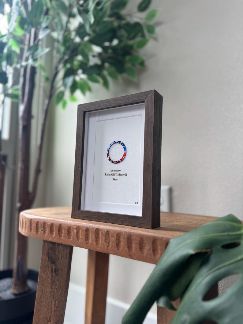 May include: A framed print featuring a watch bezel design with red, white, and blue accents. The print is set in a dark brown wooden frame with a white mat. The text "Rolex GMT Master II Pepsi" is visible.