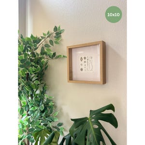 Puede incluir: Una obra de arte enmarcada con un marco de madera clara, que mide 25x25 cm. La obra presenta un dise&ntilde;o minimalista en blanco y negro sobre un fondo blanco. El marco est&aacute; montado en una pared de color claro, con vegetaci&oacute;n en primer plano.