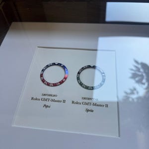 May include: Two Rolex GMT-Master II watch bezels, one with a red and blue Pepsi design, the other black and green Sprite design. The bezels are displayed in a framed presentation with model numbers and names.