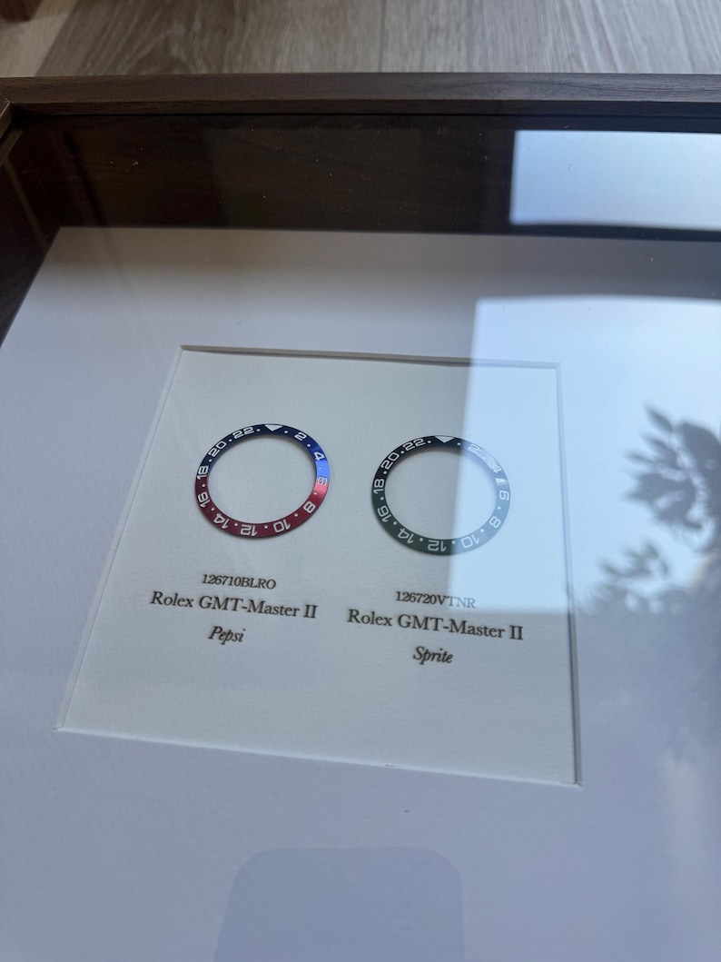 May include: Two Rolex GMT-Master II watch bezels, one with a red and blue Pepsi design, the other black and green Sprite design. The bezels are displayed in a framed presentation with model numbers and names.