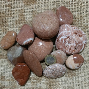 Puede incluir: Un conjunto de piedras lisas y pulidas en tonos rojos, marrones y beige. Algunas piedras tienen vetas o manchas blancas. Se incluye una moneda como referencia de tamaño. Las piedras están sobre una superficie beige texturizada.