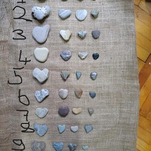 Puede incluir: Una colección de piedras en forma de corazón de varios tamaños y colores, dispuestas sobre una superficie de arpillera. Las piedras varían del gris claro al gris oscuro, con algunas que muestran variaciones blancas y de otros colores. Los números del 1 al 10 están escritos en tinta negra a la izquierda.