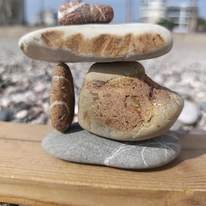 Puede incluir: Una pila equilibrada de piedras lisas y coloridas sobre una tabla de madera. La disposición incluye una piedra grande y plana de color claro, una piedra más pequeña y rayada, y una piedra de color marrón rojizo en la parte superior. El fondo muestra una playa y palmeras.