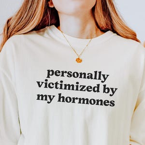 Puede incluir: Una camiseta de manga corta en color blanco roto con el texto negro "personally victimized by my hormones" impreso en una fuente sans-serif sencilla en el pecho. La prenda tiene un corte holgado con un cuello redondo cl&aacute;sico.