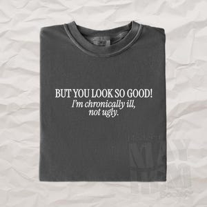 May include: A charcoal gray crew neck t-shirt featuring white text that reads, BUT YOU LOOK SO GOOD! I'm chronically ill, not ugly. The shirt has a ribbed collar and a visible brand label on the inside of the neckline, presented in a flat lay.
