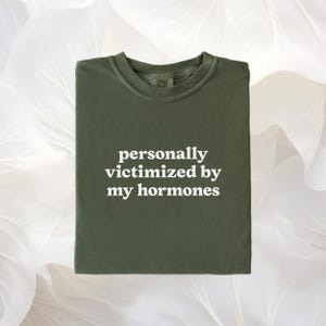 Puede incluir: Una camiseta de cuello redondo color verde oliva con el texto en blanco personally victimized by my hormones impreso en el centro. La prenda cuenta con un cuello acanalado y una etiqueta de marca visible en el interior del escote.
