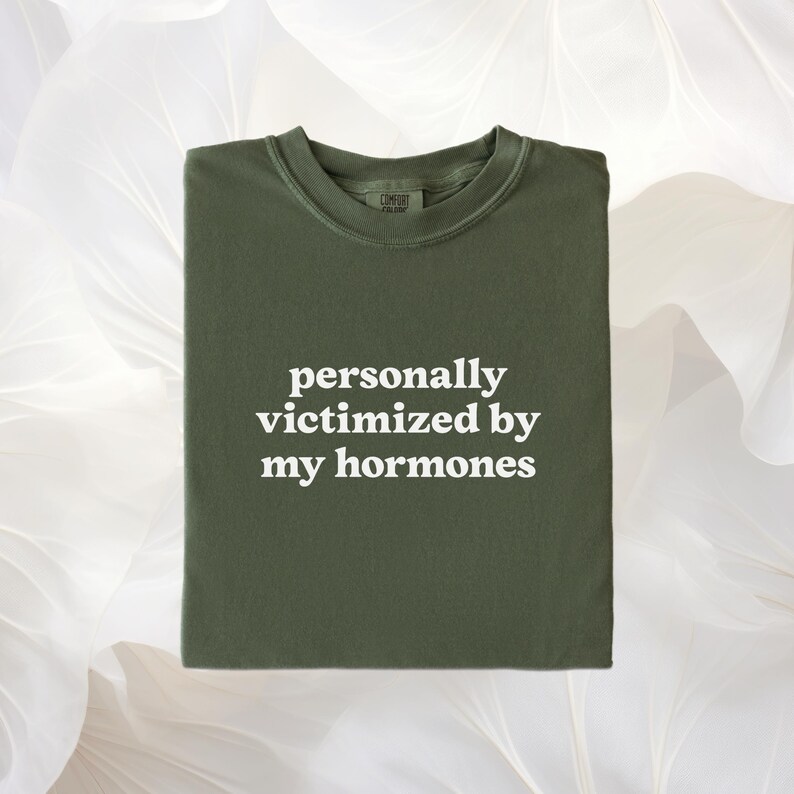 Puede incluir: Una camiseta de cuello redondo color verde oliva con el texto en blanco personally victimized by my hormones impreso en el centro. La prenda cuenta con un cuello acanalado y una etiqueta de marca visible en el interior del escote.