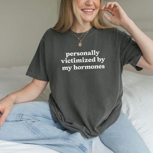Puede incluir: Una camiseta de manga corta en color gris carb&oacute;n con el texto blanco "personally victimized by my hormones" impreso en una fuente sans-serif en el pecho. La prenda tiene cuello redondo y un corte holgado y casual.
