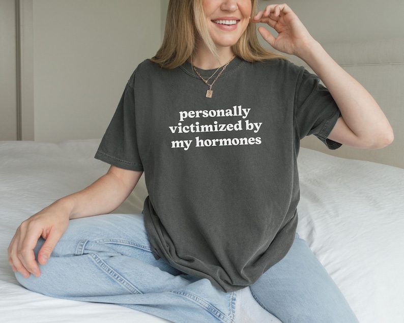 Puede incluir: Una camiseta de manga corta en color gris carb&oacute;n con el texto blanco "personally victimized by my hormones" impreso en una fuente sans-serif en el pecho. La prenda tiene cuello redondo y un corte holgado y casual.